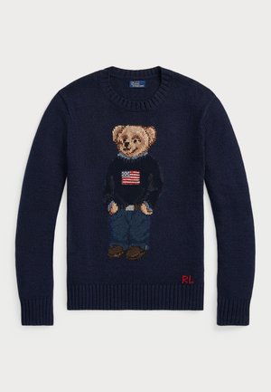 Navy knit sweater featuring an embroidered teddy bear design with a flag sweater, denim blue jeans, and brown shoes. Red logo on hem.