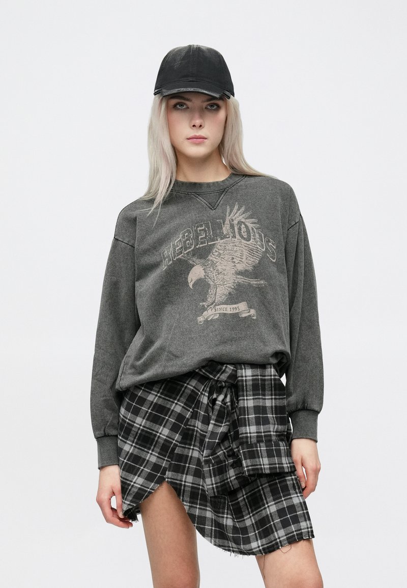 Felpa grigia con grafica di un'aquila e testo "REBELLIOUS", abbinata a una gonna a quadri in bianco e nero. Entrambi i capi presentano un fit rilassato.