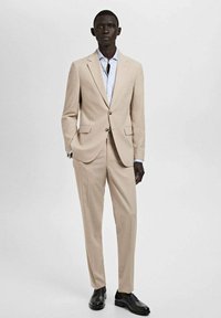 Beige suit made of textured fabric, featuring a two-button blazer, flat-front trousers, and a light blue shirt underneath. Black shoes.