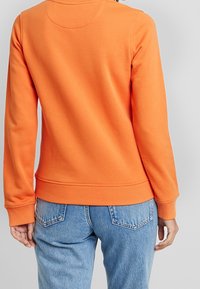 Felpa arancione con maniche lunghe e polsini a costine, abbinata a jeans azzurri. Tessuto morbido, vestibilità casual e vita a banda.