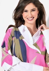 V-Ausschnitt Pullover mit auffälligen Blumenmustern in Pink, Marineblau und Oliv auf einem strukturierten Stoff, über einem weißen Hemd mit Kragen getragen.