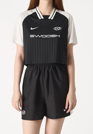 Vrouw draagt een zwart cropped sportshirt met witte mouwen en bijpassende zwarte shorts, beide met witte logo's en een atletisch ontwerp.