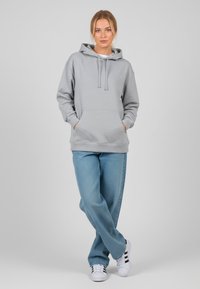 Felpa grigia con tasca a marsupio, cappuccio con cordino, vestibilità comoda. Abbinata a jeans azzurri e sneakers bianche con strisce nere.