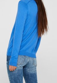 Maglione blu a maglia con maniche lunghe, polsini e orlo a costine, abbinato a jeans di denim scoloriti. La texture appare morbida e liscia.