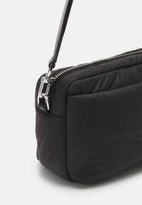 Borsa puffer nera con una texture liscia e imbottita. Presenta una chiusura con zip argentata e una tracolla in pelle regolabile e elegante per il trasporto.