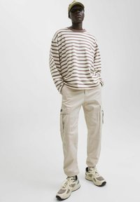 Langärmeliges gestreiftes Hemd in Beige und Braun, kombiniert mit beigen Cargohosen mit Seitentaschen; dazu beige Sneaker und eine Kappe.