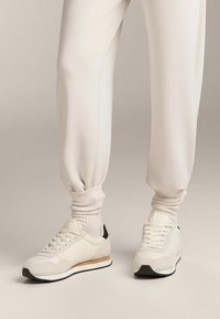 Krämfärgade sportskor i texturerat tyg med svart hälaccent och vit sula. Kombinerade med ljusa, ribbade strumpor och joggingbyxor.