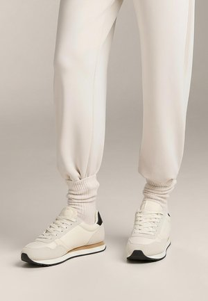 Kremowe sportowe trampki z teksturowaną tkaniną, czarnym akcentem na pięcie i białą podeszwą. W zestawie z jasnymi, prążkowanymi skarpetami i dresami.