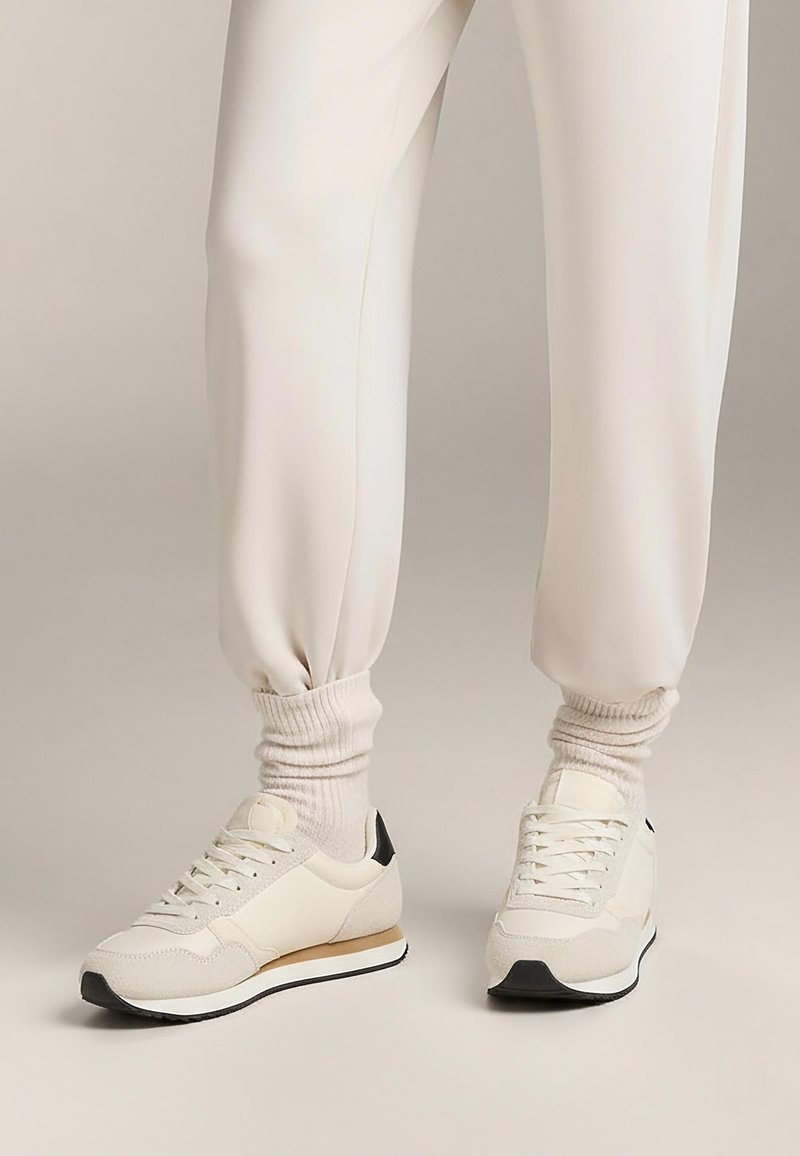 Krämfärgade sportskor i texturerat tyg med svart hälaccent och vit sula. Kombinerade med ljusa, ribbade strumpor och joggingbyxor.