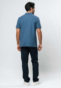 Mies seisoo selin päin, yllään sininen lyhythihainen poolopaita, tummat farkut ja valkoiset tennarit, yksinkertaisen vaalean taustan edessä.