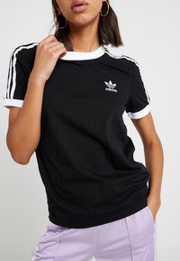 Černé tričko s bílým zdobením a třemi bílými pruhy na rukávech. Na hrudi je malé bílé logo Adidas. Krátký rukáv, bavlněný materiál.