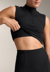 Top nero senza maniche con colletto a metà zip, tessuto elasticizzato e vita arricciata, abbinato a pantaloni neri a vita alta con dettagli testurizzati.
