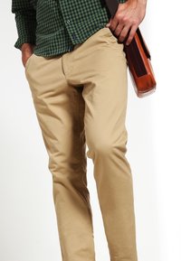 Pantalon chino beige en coton à la texture lisse, coupe slim, porté avec une chemise à carreaux verte et noire, et un sac messager en cuir.