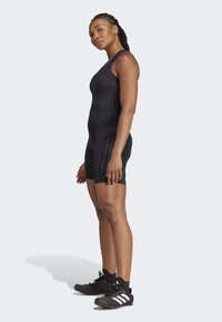 Black athletic bodysuit with a fitted design, knee-length shorts, and subtle striping detail, paired with black athletic shoes featuring white stripes.