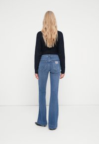 Jeans de denim azul com boca de sino e cintura média, apresentando um design de bolso traseiro e costura amarela, combinados com um suéter de malha preto.