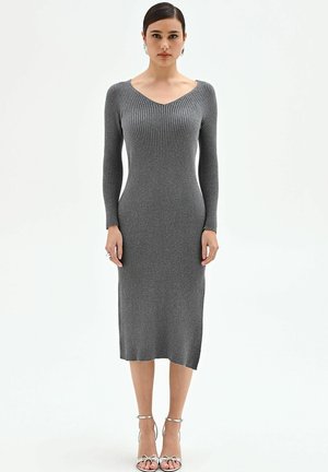 Donna che indossa un abito midi grigio a costine con maniche lunghe e sandali argentati con tacco alto, in piedi davanti a uno sfondo bianco.