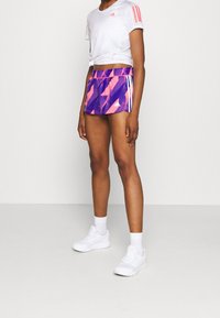 Vit kort t-shirt med rosa ränder på ärmarna, tillsammans med lila och rosa geometriska shorts. Vita sportskor och strumpor.