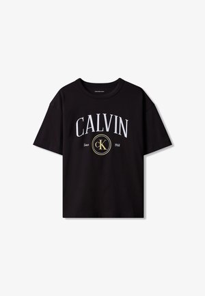 Chłopiec ubrany w czarną koszulkę Calvin Klein, niebieskie dżinsy, białe trampki, z niebieską kurtką jeansową zawiązaną wokół talii, stojący na szarym tle.