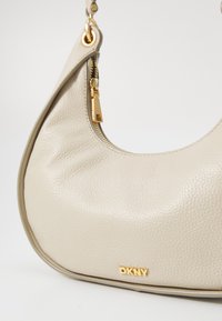 Borsa a tracolla in pelle beige con design curvo, superficie testurizzata, accessori dorati, chiusura con zip e un evidente dettaglio del logo DKNY.