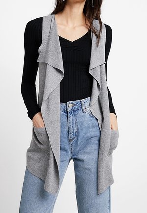 Gilet grigio chiaro aperto realizzato in morbido tessuto, design a strati con colletto drappeggiato e due tasche frontali, indossato sopra un top nero a coste e jeans blu.