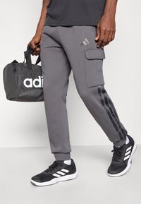 Pantalones de chándal grises con bolsillo cargo lateral, detalle de raya negra en las piernas, combinados con zapatillas deportivas negras y una bolsa de deporte gris con marca blanca.