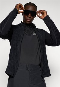 Vest negru din fleece cu țesătură texturată, suprapus peste un top termal negru. Modelul poartă ochelari de soare închiși la culoare, prezentând un aspect elegant și modern.