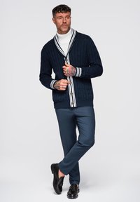 Cardigan navy a maglia cable-knit con dettagli a strisce bianche e bottoni, abbinato a pantaloni scuri slim-fit e mocassini neri.