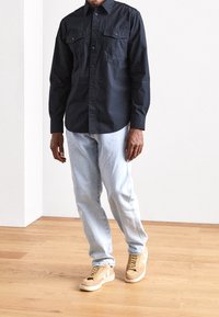 Camisa de botón azul oscuro con bolsillos en el pecho, pantalones vaqueros azul claro y zapatillas color arena con acentos blancos, usadas por una persona de pie.