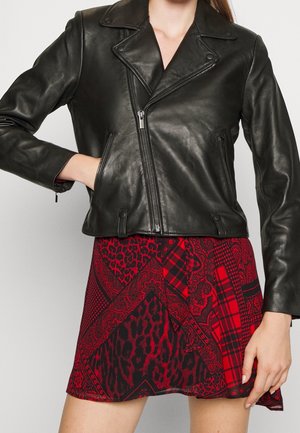 Woman wearing a black leather zip-up jacket with collar over a red and black patterned skirt, hand in jacket pocket.