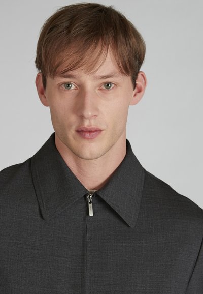 Veste col noir en gris foncé avec un détail zippé, présentant un tissu texturé et un design épuré et structuré.