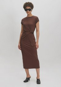 Braunes, tailliertes Midi-Kleid mit kurzen Ärmeln und seitlichem Bindedetail. Glatter, dehnbarer Stoff. Kombiniert mit schwarzen Mules und übergroßen Sonnenbrillen.