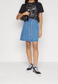 Svart t-shirt med vit linjeteckning av ett ansikte, kombinerad med en blå denim A-linje kjol med framknappar, och svarta högknäppta sneakers.