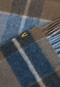 Écharpe en plaid en laine présentant des nuances de bleu et de marron, avec une texture douce et des bords à franges. Comprend une petite étiquette noire avec un logo jaune.