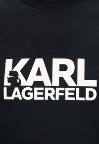 Μαύρο ύφασμα με έντονο λευκό κείμενο «KARL LAGERFELD», με μια στυλιζαρισμένη σιλουέτα ατόμου με γυαλιά ηλίου και κότσο μέσα στο γράμμα K.