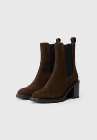 Botas de tobillo de ante marrón con paneles laterales elásticos negros, punta redondeada y tacón cuadrado. La superficie texturizada añade profundidad al diseño.
