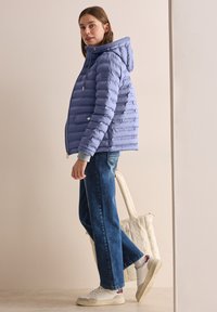 Ljuslila puffjacka med horisontell quiltning, justerbar huva och grå manchetter. Ett par blå jeans och vita sneakers, håller en krämfärgad tote-väska.