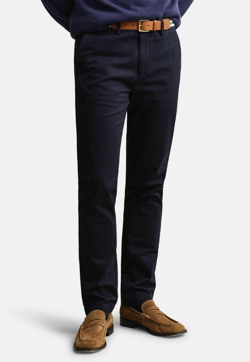 Homme portant un pantalon slim bleu marine foncé avec une ceinture en cuir marron et des mocassins en daim marron assortis sur fond blanc.