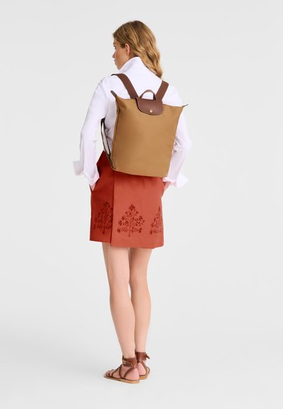 Brown backpack with a flap and button closure, featuring brown straps. Worn with an orange embroidered skirt and white long-sleeve shirt.