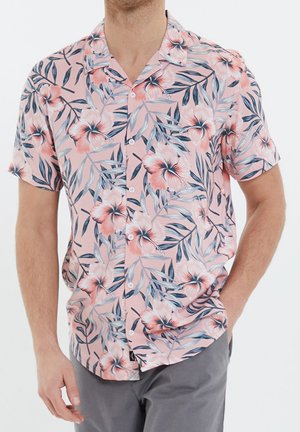 Homme portant une chemise rose à manches courtes avec un grand motif floral et feuillu dans des tons bleus et blancs.