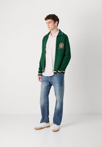 Groen gebreide cardigan met gestreepte kraag en embleem, gedragen over een wit en lichtblauw gestreept shirt, gecombineerd met blauwe jeans en witte sneakers.