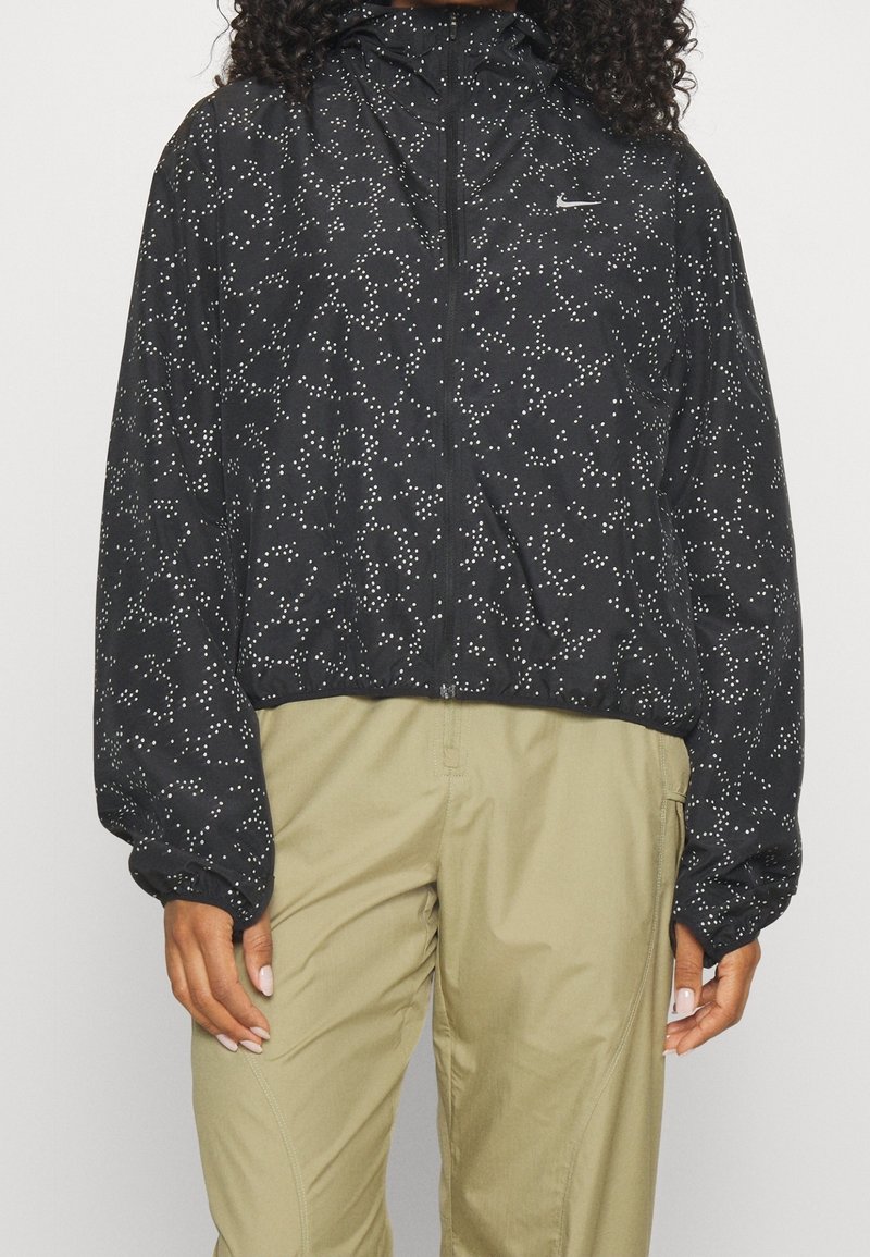 Veste noire zippée avec un motif d'étoiles en points blancs, portée avec un pantalon beige, montrée du cou jusqu'à mi-cuisse sur une personne debout.