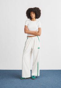 Calças de fato de treino brancas, largas, com riscas verdes nas laterais e logótipo da Adidas. Combinadas com um top branco justo de manga curta e ténis verdes.