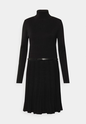 Robe en tricot noire à manches longues avec col roulé et jupe plissée, accessoirisée d'une ceinture noire fine à la taille.