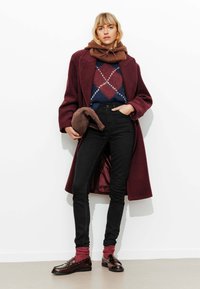 Manteau bordeaux sur un pull à motifs, jeans noirs et mocassins marron avec des chaussettes rouges. Texture lisse, design structuré, style décontracté.