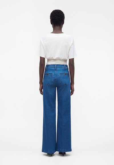 Personne portant un haut blanc à manches courtes et un jean bleu taille haute à jambes larges avec poches arrière boutonnées, debout de dos sur fond blanc.
