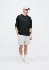 Μαύρο βαμβακερό μπλουζάκι με κόκκινο γραφικό στοιχείο, γκρι φλις σορτς, γκρι καπέλο μπέιζμπολ, λευκά αθλητικά παπούτσια και ανοιχτό γκρι κάλτσες. Καθημερινή αθλητική ενδυμασία.