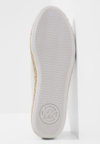 Semelle d'une chaussure blanche avec un motif ondulé et un logo circulaire "MK" près du talon, inclinée contre un mur blanc.