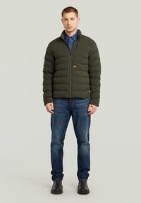 Giacca piumino verde scuro con zip frontale, dotata di colletto arrotondato e motivo trapuntato, abbinata a jeans in denim blu e stivali neri.