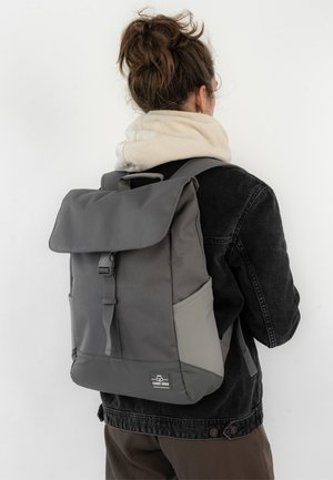 Grey backpack with a flap closure, made of durable fabric. Features a front buckle, side pockets, and a textured base for stability.