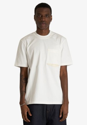 Uomo che indossa una semplice T-shirt bianca a maniche corte con taschino sul petto e jeans scuri, in piedi davanti a uno sfondo chiaro.